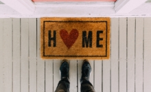 a welcome mat on the ground in front of a pink door that says Home with the O being a red heart