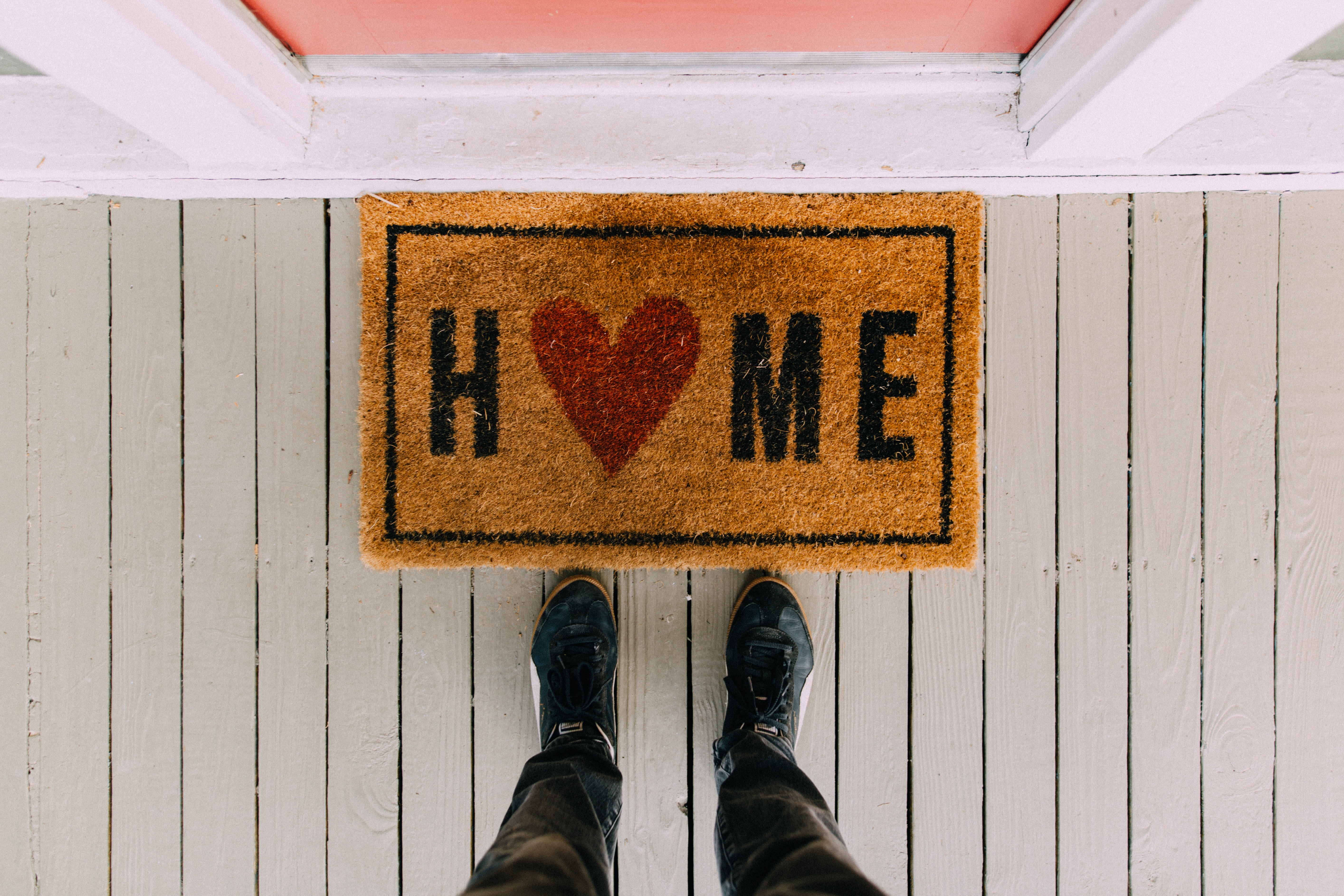 a welcome mat on the ground in front of a pink door that says Home with the O being a red heart