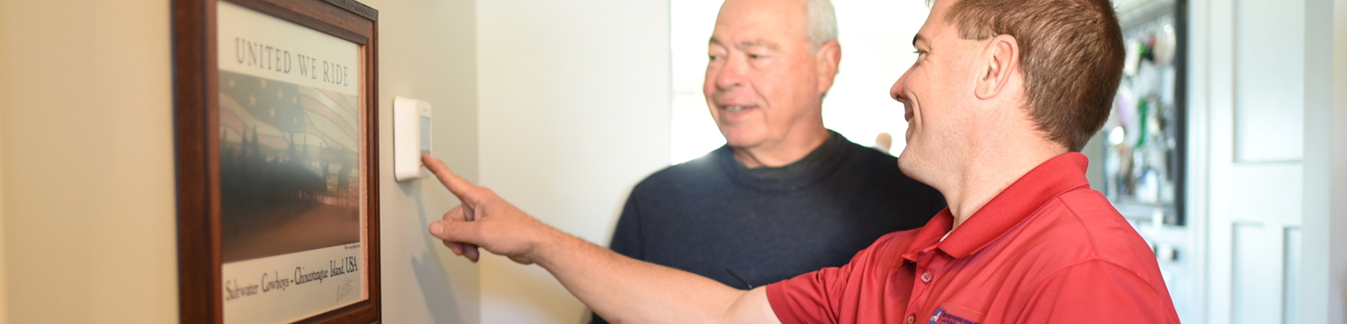 HVAC technician in a red polo shirt installing a smart thermostat on a home hallway wall.